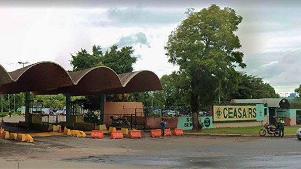 Vista da rua de frente para p&oacute;rtico de entrada de autom&oacute;veis da CEASA/RS, com um painel ao lado direto com a marca e nome CEASA RS
