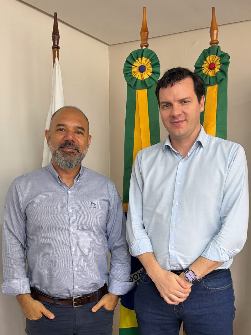 O representante da CEASA/RS junto ao representante da AGAS, posam para foto em frente a bandeiras com fitas verdes e amarelas. Ambos usam camisas sociais claras e sorriem levemente para a c&acirc;mera