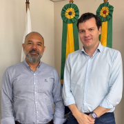O representante da CEASA/RS junto ao representante da AGAS, posam para foto em frente a bandeiras com fitas verdes e amarelas. Ambos usam camisas sociais claras e sorriem levemente para a c&acirc;mera